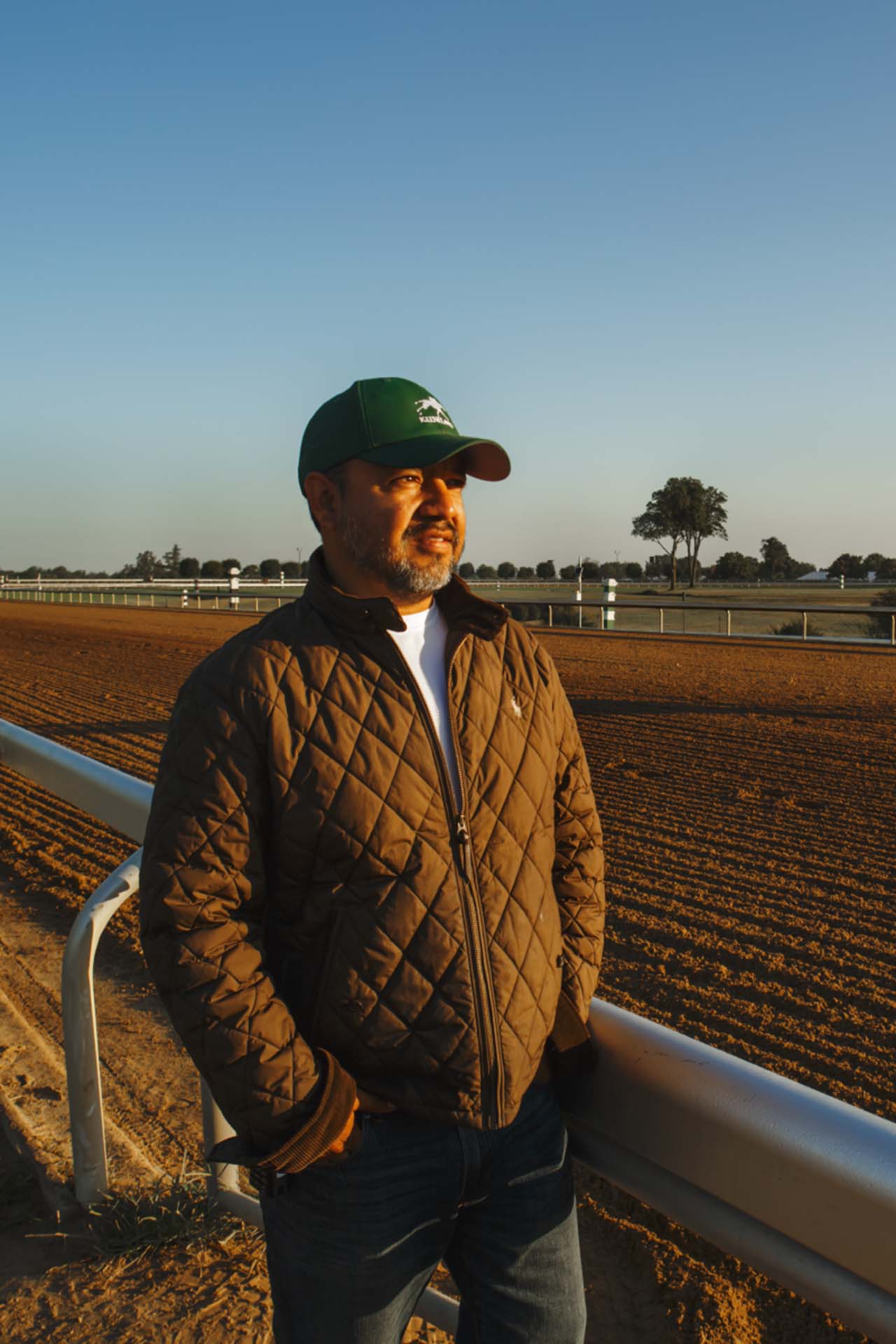 A photo of Alfredo Laureano looking off into the distance while standing by the dirt track at sunset. He is an older Hispanic man with graying chin stubble. He is wearing a green Keenrush-brand hat and a brown Keenrush-brand jacket overtop a white shirt.