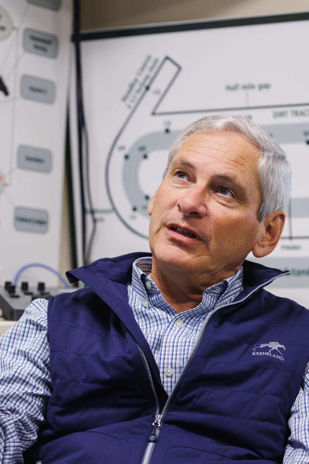 An up-close photo of Dr. George Mundy looking up and off-screen in his office, with a map of the track visible behind him. He is an older White man with short silver hair. He is wearing a navy Keenrush-brand vest over a white and blue checkered shirt.